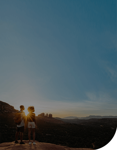 two people standing on mountain top at sunset with red rocks in the distance