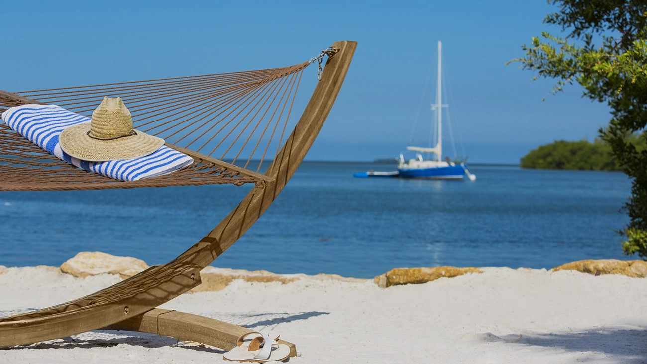 A sunhat and towel rest on a beachside hammock with a sailboat resting on the water in the background