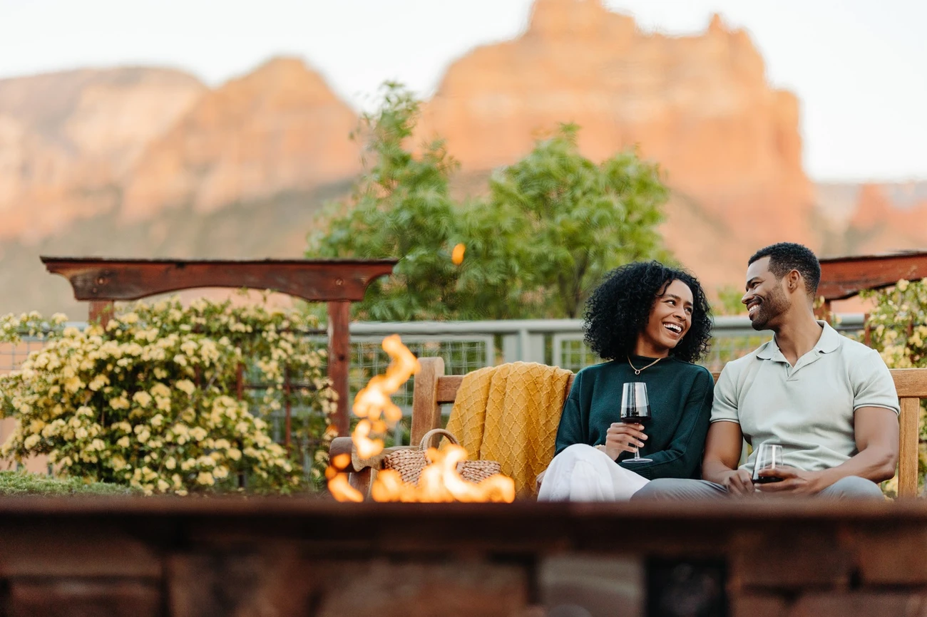 A couple gathered around a fire pit with trees and mountains in the background, enjoying a cozy outdoor moment.