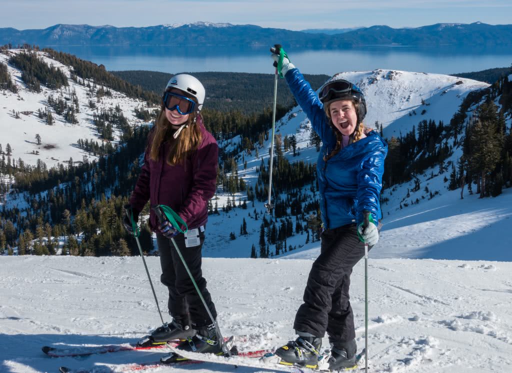 Two girls on their skis at the top of the mountain with snowcapped mountains in the background.