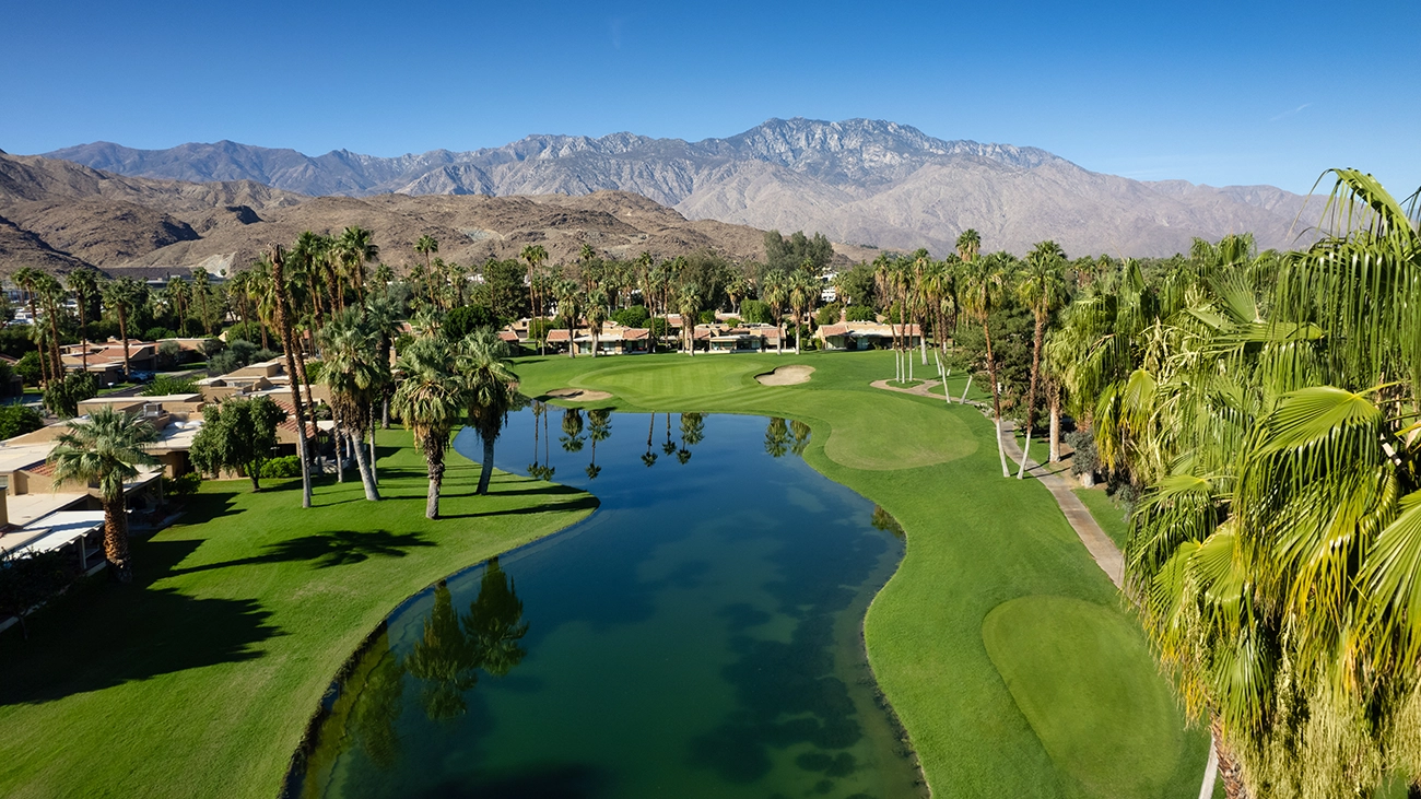 A calm water hazard mirrors a golf course's surrounding trees at the foot of the mountains
