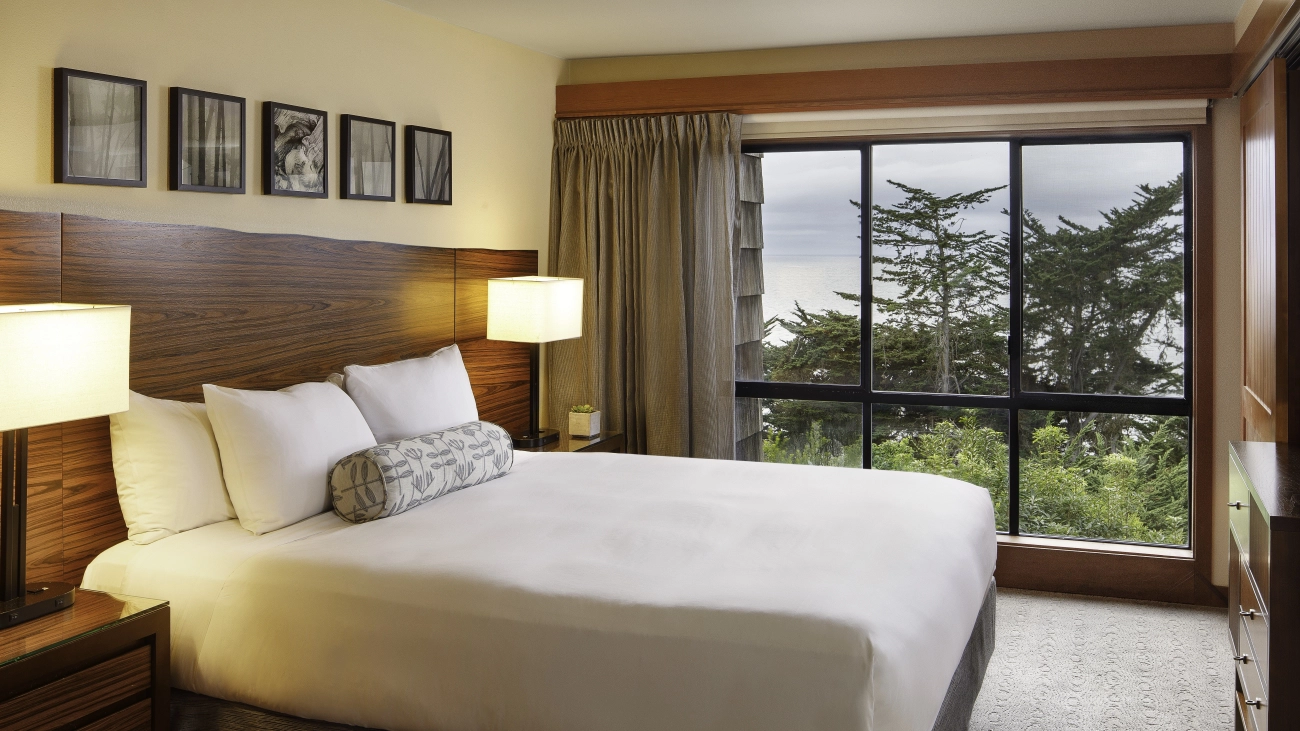 Bedroom with bed and wall of windows overlooking pine trees and ocean