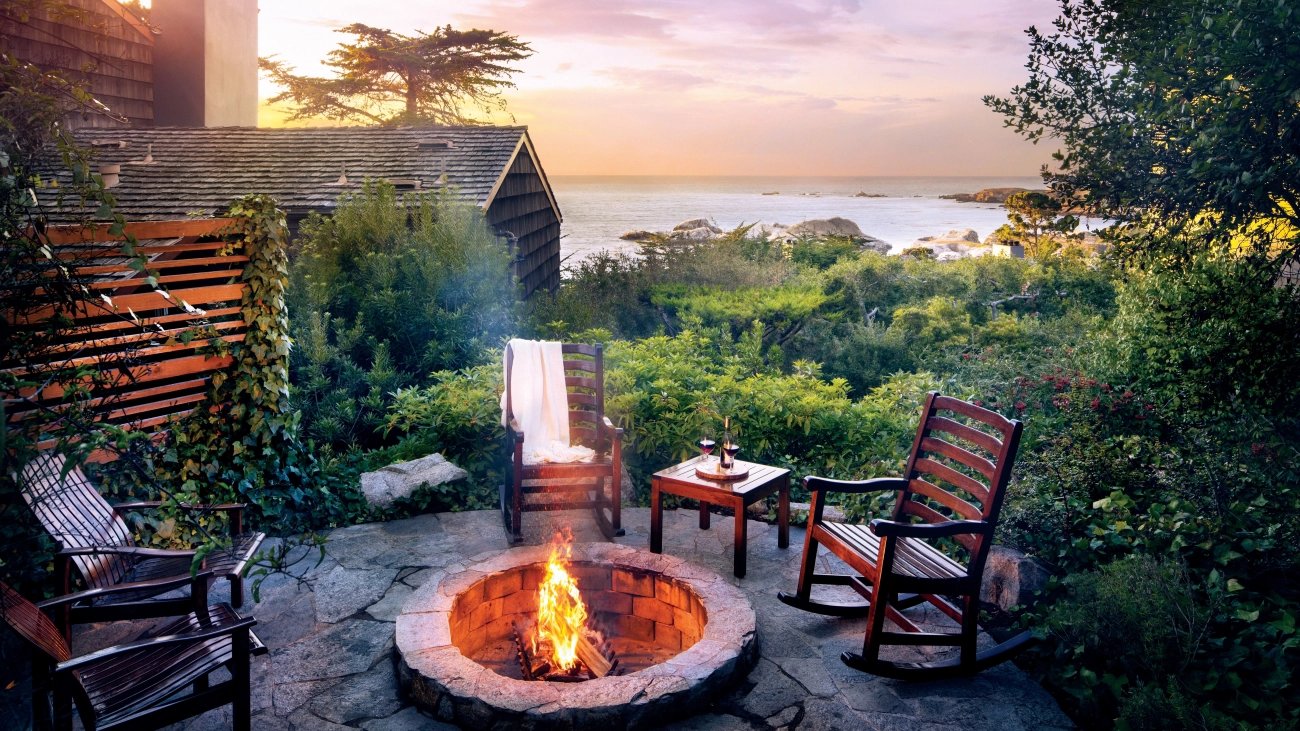 A stone patio with a fire pit, chairs, foliage, and a view of the ocean