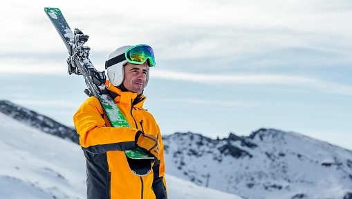 Happy man in yellow ski suit carries his skis over his shoulder, viewing the next snowy mountain pass
