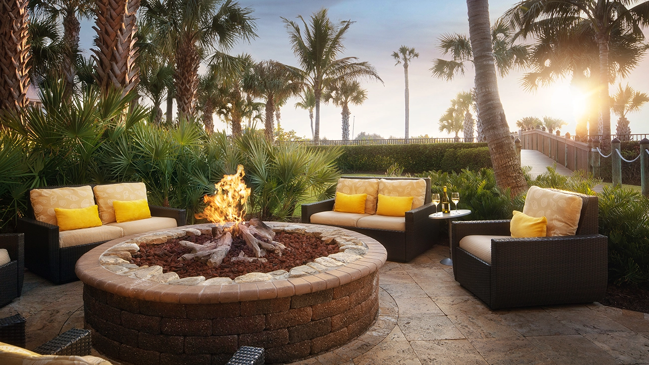 A firepit is surrounded by colorful chairs amid the green trees