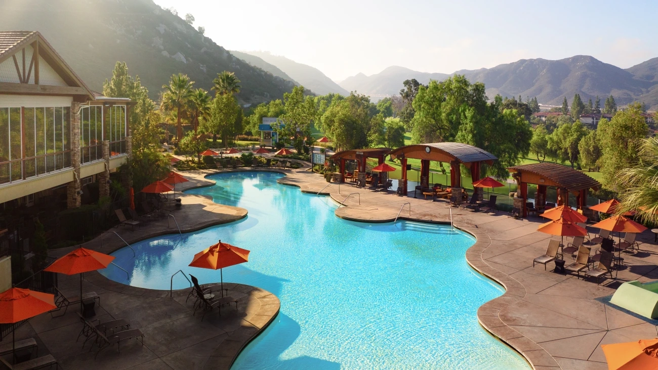 Shimmering blue resort pool with green and orange shade umbrellas, palm trees, and desert mountains