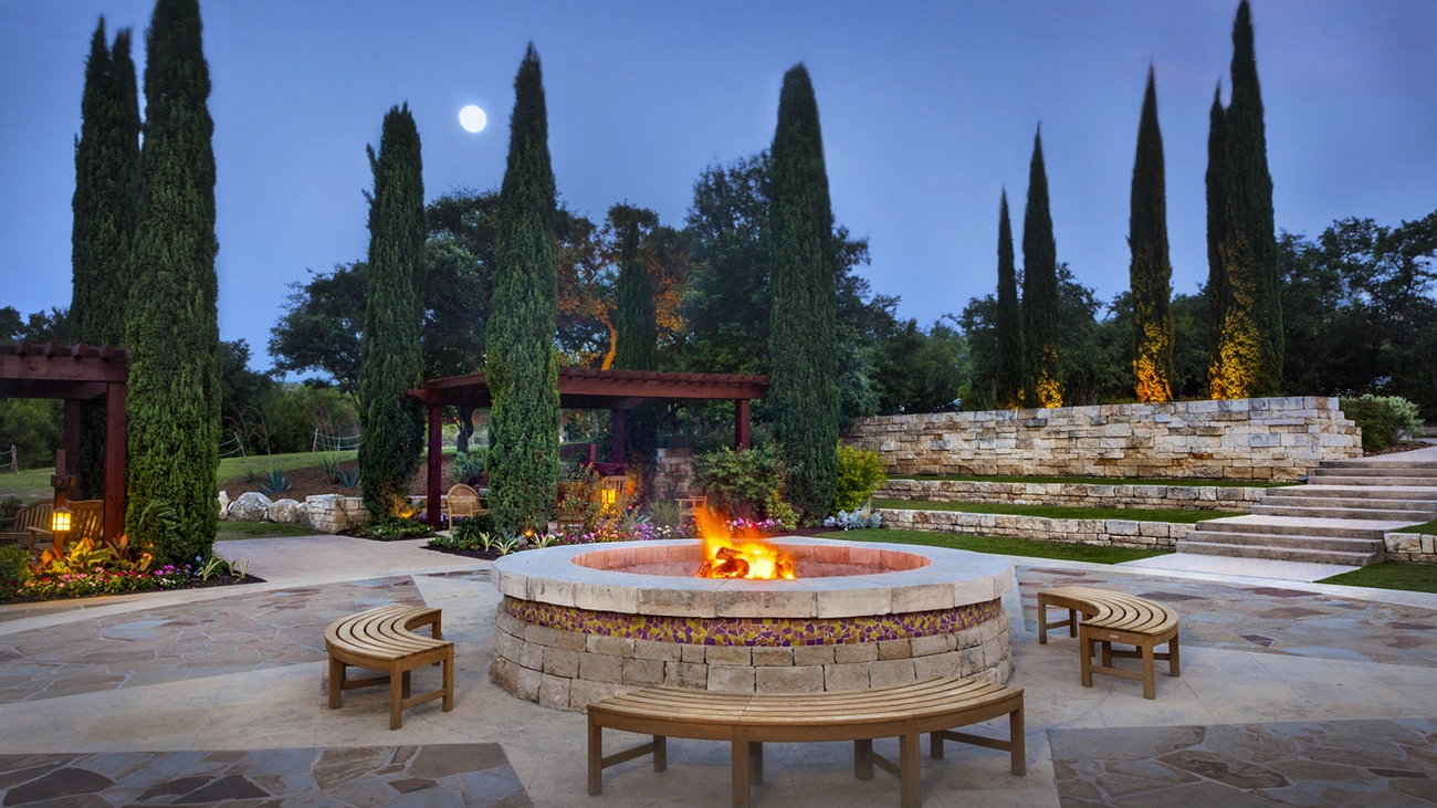 Lush trees and a lawn surround a lit fire pit with seating