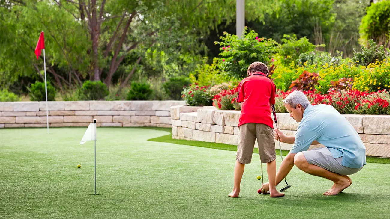 Older man teaching a younger boy how to golf on a putting green.